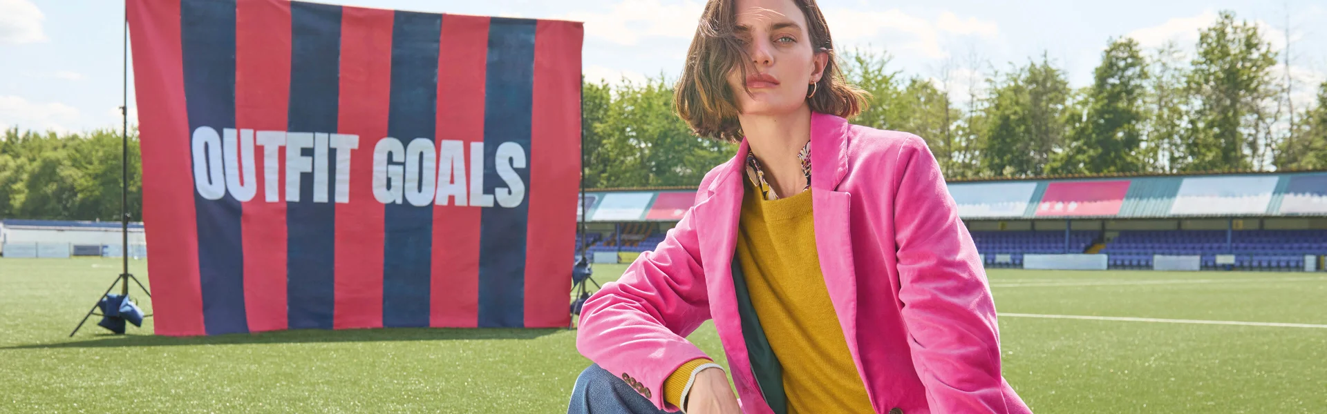 Femme en blazer rose et pull jaune, derrière elle un drapeau rayé avec l'inscription, Outfit Goals
